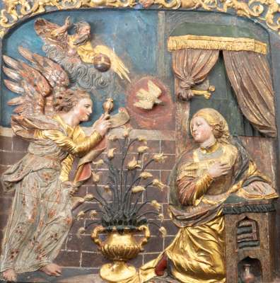 <p>Met de voorstelling van de Annunciatie. De aartsengel Gabriel, met vleugels, staat aan de linkerzijde en reikt naar Maria, die de boodschap in ontvangst neemt. Boven Maria bevindt zich een duif die de Heilige Geest symboliseert.</p>