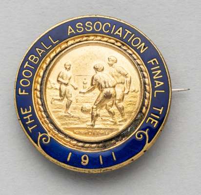 The Football Association final tie 1911 lapel badge,
 obverse with footballers in relief surrounded by blue enamel lettering THE FOOTBALL ASSOCIATION 